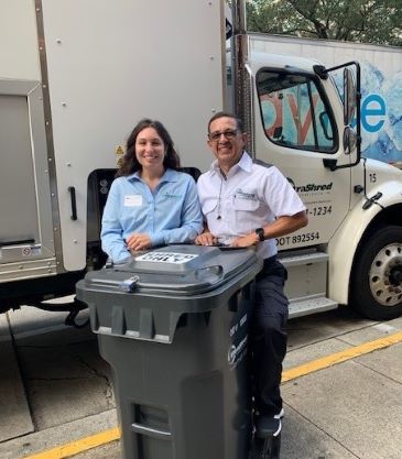 Two people standing with a grey collection container in front of an UltraShred mobile shredding truck
